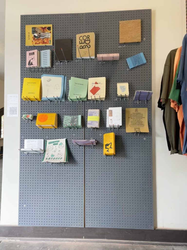 Image: Forward-facing view of the gray pegboard showcasing zines for sale by Aguilar and Knecht. They vary in color, size, and shape. Image by Nadia John.