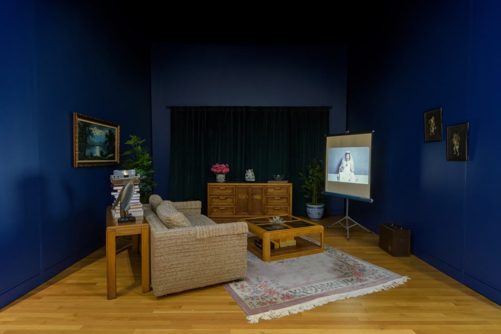 (Right) Strange You Never Knew, 2025. Photograph of a living room from the exhibition Strange You Never Knew. The room has framed art hanging from the dark blue walls, a couch faces a projector screen, and other pieces of furniture fill the space. Photo credit: Charles Edwards. Images courtesy of the University of Michigan Museum of Art.