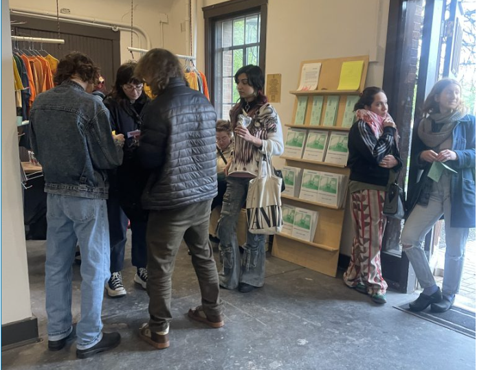 Image: A crowd stands around the entrance of Comfort Station and the store installed by Aguilar and Knecht, which includes a collection of their zines and homemade tee's which each have cheeky phrases stamped upon them. On the wall of Comfort Station near the entrance doorway, exhibition brochures can be seen lined on a tiered wooden shelf. Image by Nadia John.