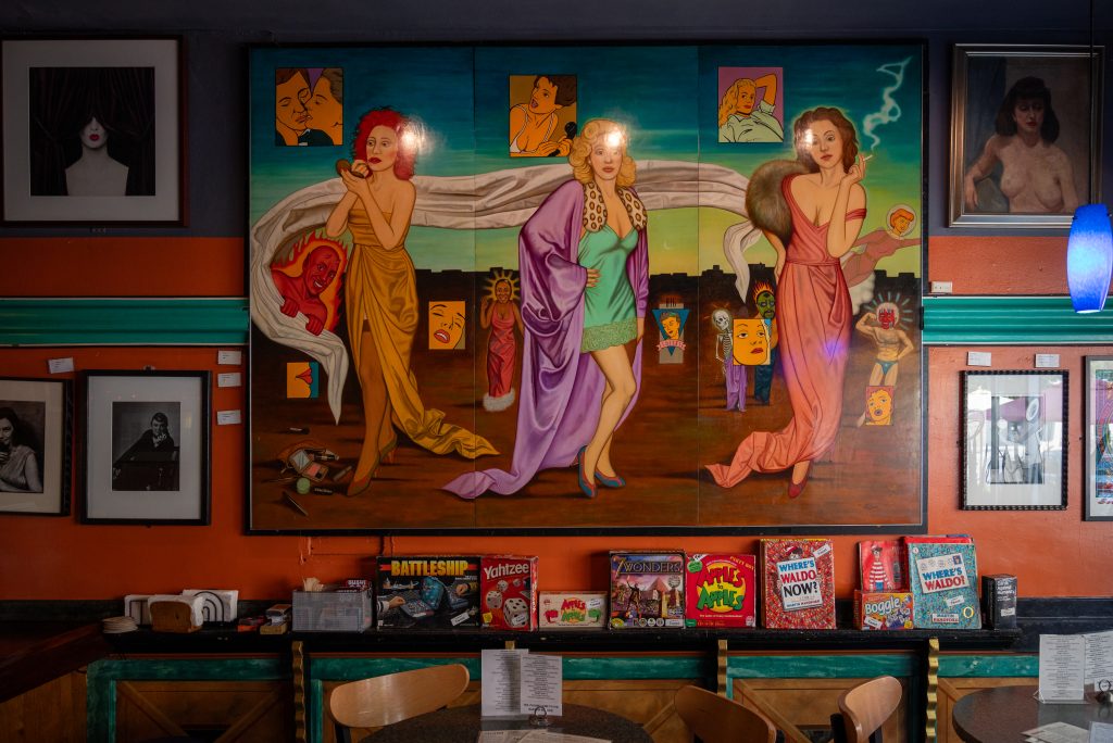 Image: View of a large painting positioned above a shelf full of board games and wood tables and chairs. The painting is from the Chicago Imagist tradition. It features a triptych of stylish women in various stages and experiences in their lives. Behind one of the woman is a smiling devil hanging on a fabric that started flowing out from one of the homes in the row of building behind the women. Image by Tonal Simmons.