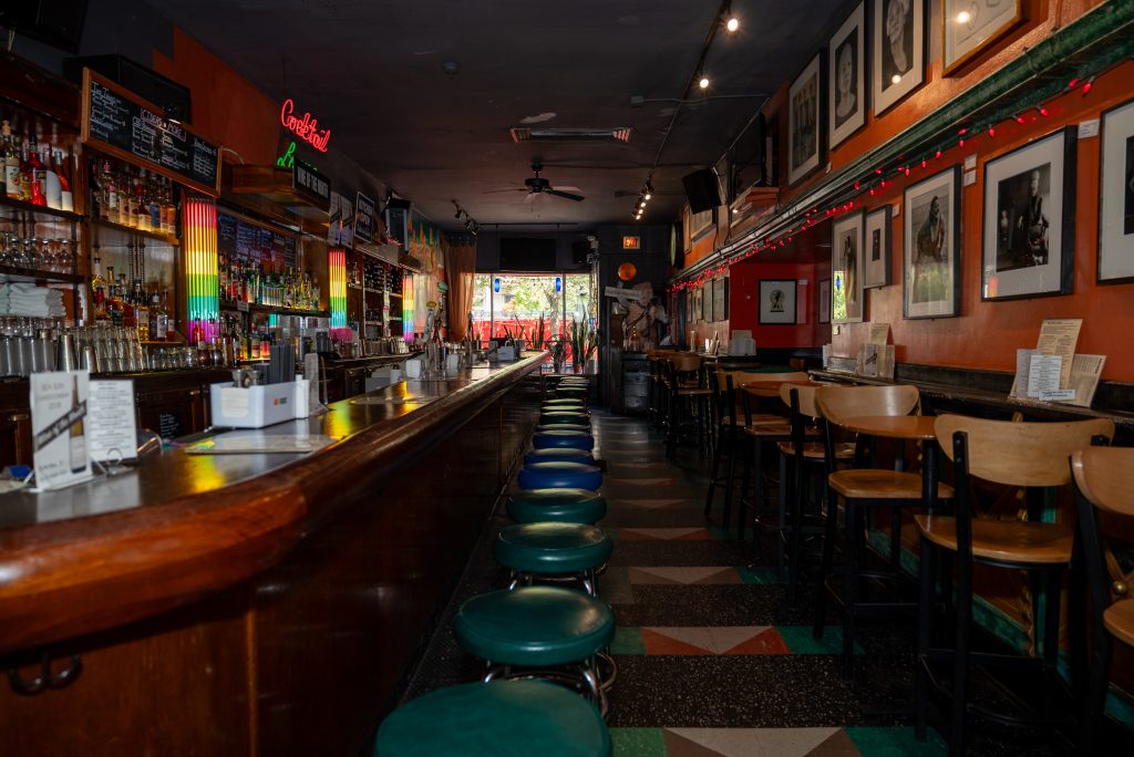 Image: View of Big Chicks bar facing from the back of the bar towards the front. A row of green cushioned bar stools are positioned in front of the bar and wood seats with tables are positioned on the opposite wall under framed art. Image by Tonal Simmons.