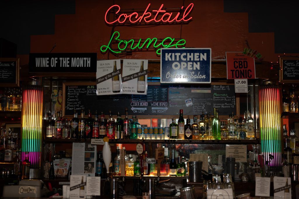 Image: Close up of the bar at Big Chicks, which features the words "Cocktail Lounge" in red and green neon lights in the center above the bar. Below it, rainbow color-blocked pillars stand on each side and bottles of drinks cover the shelves. Image by Tonal Simmons.