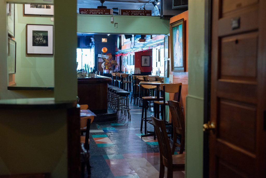 Image: A view of the bar from a far back corner near a bathroom door. Visible in this image is the colorful tiling under the bar stools and chairs. Bright sunlight shines through the front window to illuminate the space. Image by Tonal Simmons.