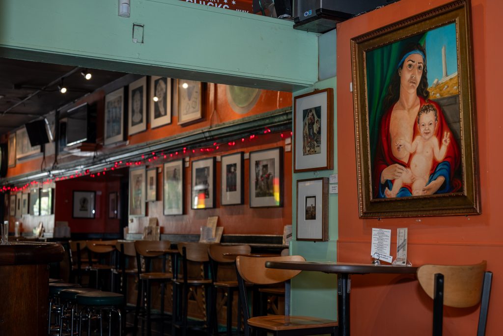 Images: (Above) View of the bar facing the rows of tables. The walls are painted orange, except for where there are partitions where its painted teal. Framed art can be seen lining the walls. The closest image is a figure of a mother and child. The child is pinching their mother's nipple and making a peace sign with their other hand. (Images 1-6 below) Fire’s art collection continues to adorn the walls. Various framed art hang on the walls at Big Chicks. Some walls are orange, some green, and some pale yellow. Images by Tonal Simmons.