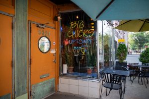 Image: Outside view of Big Chicks. Chairs for outdoor seating during morning brunch are shown as well as as a view down Broadway Ave. The front door of Big Chicks is orange. On the window outside the bar a "Big Chicks" sign, which includes three mini drinks, is done in yellow, while the words "Uptown" and "Bar" are done in blue neon lights. Image by Tonal Simmons.