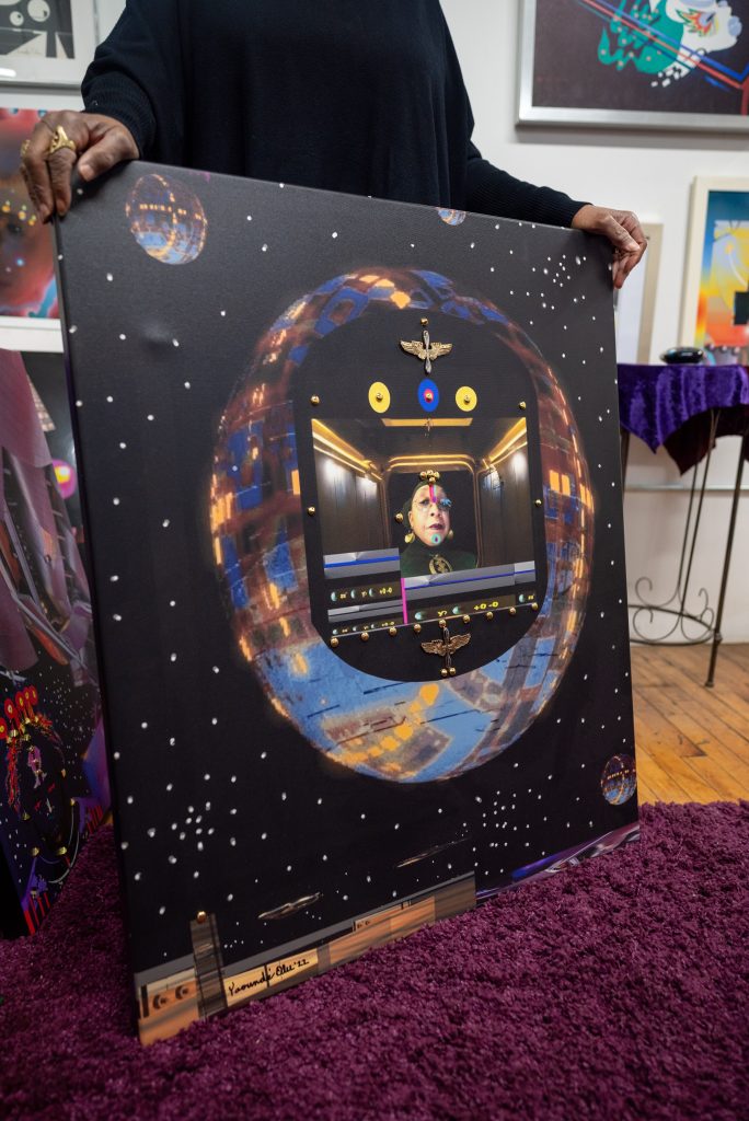 Image: (Closeup) Olu holds up a recent work. She is in the center of a globe shaped spaceship. Eagle pendants are placed around the opening of the spaceship. Image by Tonal SImmons.