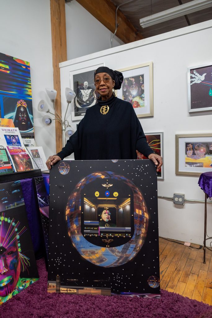 Image: Olu holds up a recent work. She is in the center of a globe shaped spaceship. Eagle pendants are placed around the opening of the spaceship. Image by Tonal SImmons.