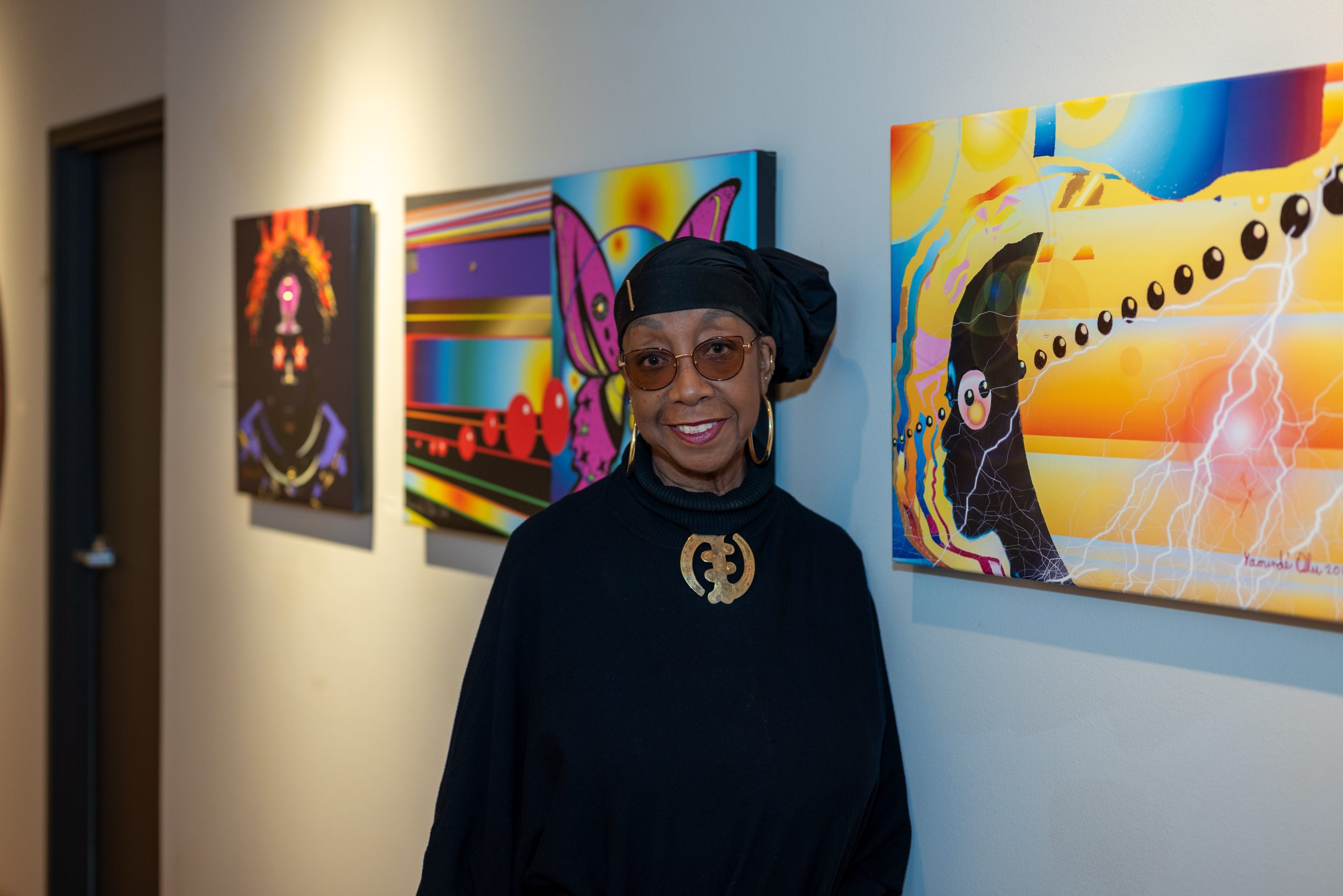 Image: Yaoundé Olu, dressed in all black with handmade gold jewlery on her fingers and neck, stands smiling in front of the work displayed on the wall in her showroom. Image by Tonal Simmons.