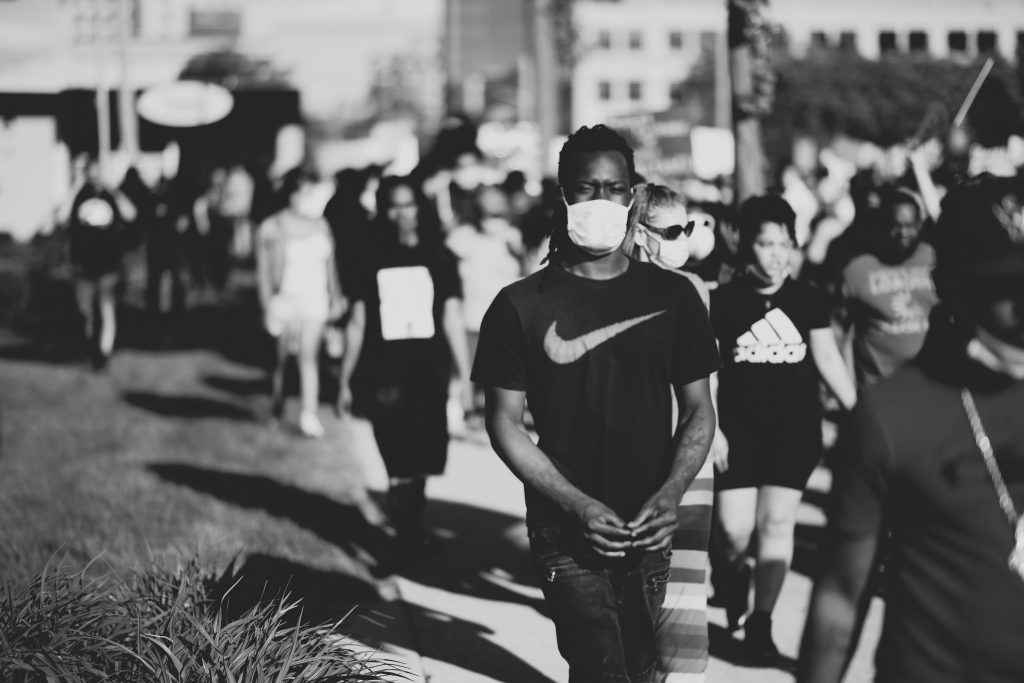 Image: “Unrest” by Xen. A black and white photograph of a crowd of people during a protest in Rockford. The person in the foreground wears a Nike shirt and a mask. Courtesy of Belt Publishing.