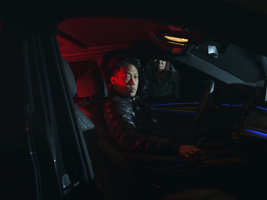 Image: Untitled (Mom and Jake), 2023, Archival pigment print. A person sits in the driver seat of a car and faces the camera. Red and white light illuminates their face. A second person's face can be seen outside the car through the slightly-open window. Image courtesy of artist.