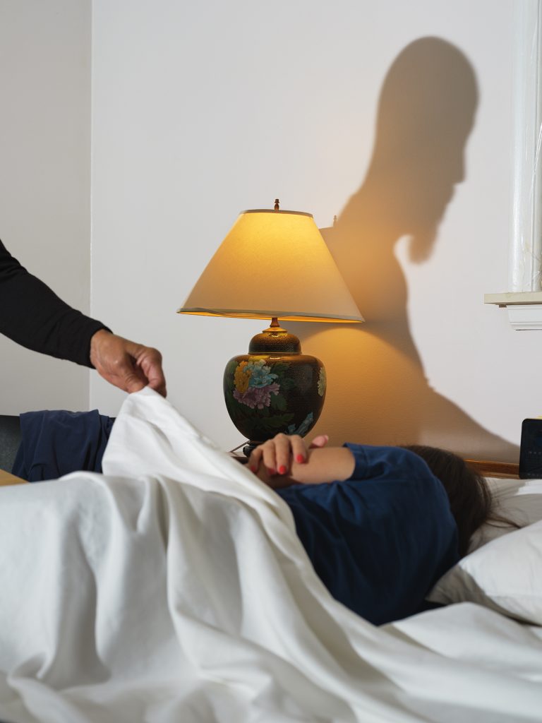 (Right) Untitled (Bedtime), 2021, Archival pigment print. One person is lying in bed, facing away from the camera while a second person, whose silhouette is cast in shadow in the wall behind the bed, pulls a white sheet over them. Images courtesy of the artist.
