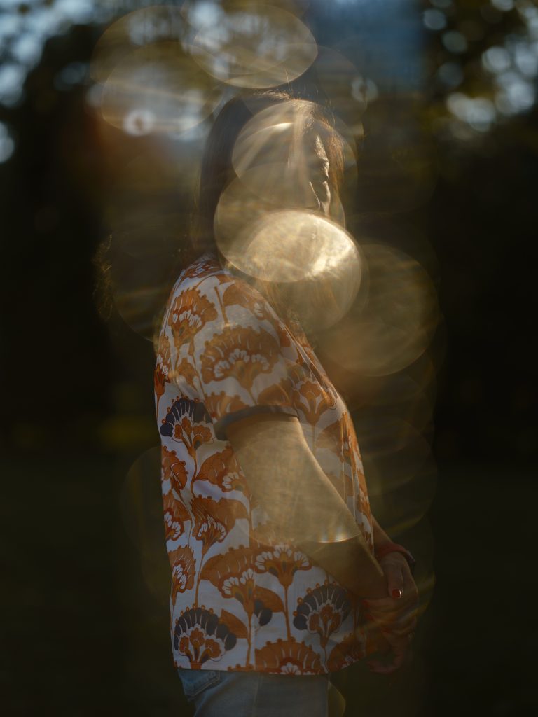 Image: Untitled (Mom), 2021, Archival pigment print. A photograph of a person standing in profile but their face is looking at the camera. Lens flare distort the image so the person is not fully in focus. They wear a white tee shirt with orange and blue detail. Image courtesy of the artist.