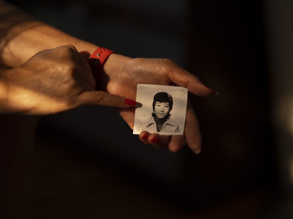Image: Untitled (Picture of Mom), 2021, Archival pigment print. Against a black background, someone's hand holds a small black and white portrait of a person with short black hair and a collar shirt. A pinky with a long red nail, points at the black and white image. Image courtesy of the artist.