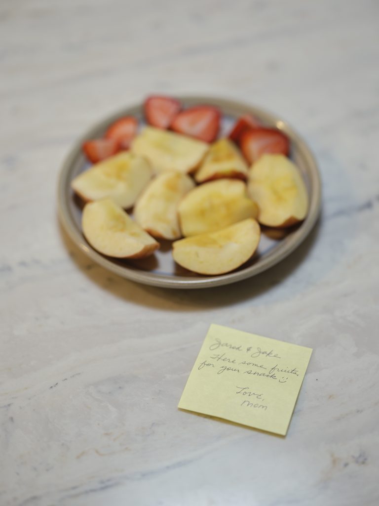 Untitled (Snack), 2021, Archival pigment print. (Left) A yellow post it note with words written in smooth, cursive writing is placed in front of ceramic plate full of browning apple slices and cut strawberries. The words "love mom" can be made out on the note.