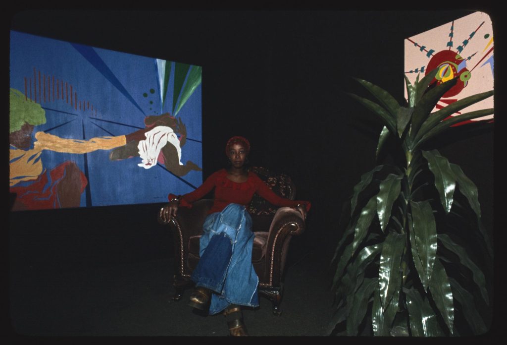 Image: Yaoundé Olu,  seated in a chair at her Oşun gallery, 2541 E. 75th St., Chicago, Illinois, in front of her work. Image by Jonas Dovydenas for the Library of Congress' Chicago Ethnic Arts Project. 