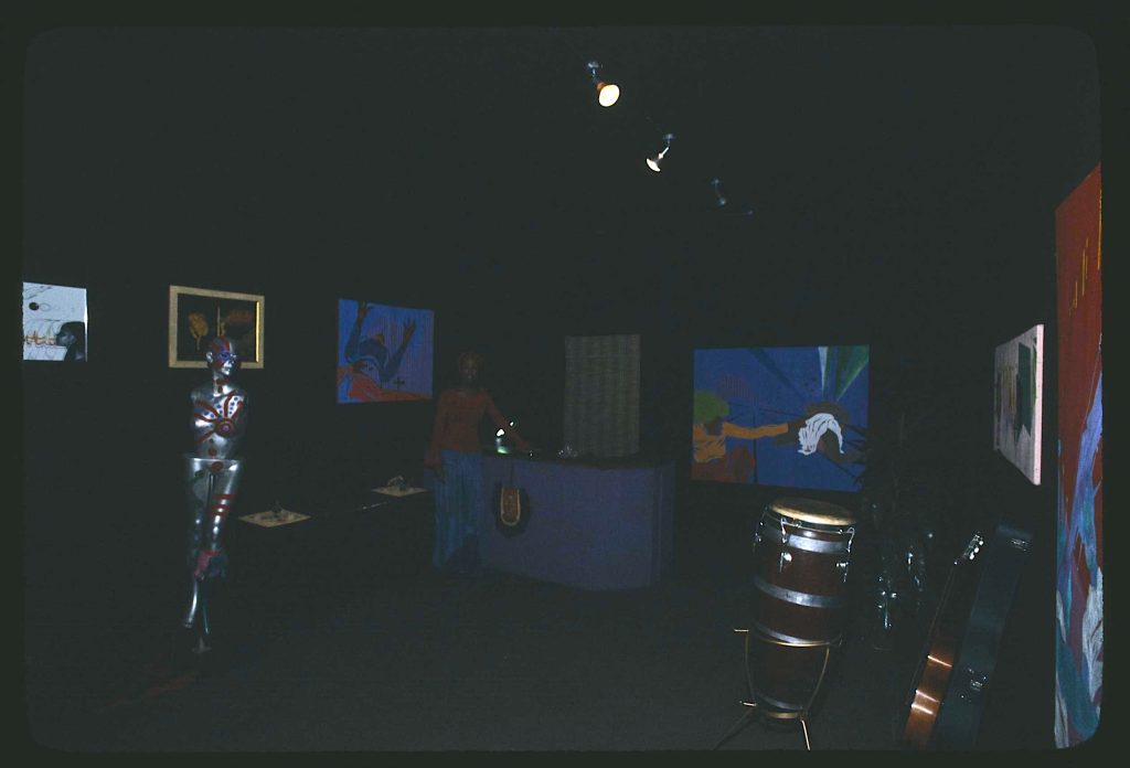 Image: Yaoundé Olu, uniphysicist, at her Osun Studio, 2541 E. 75th St., Chicago, Illinois, standing by her reception desk in her exhibition space. An exhibition of her work, "Galaxy Council" is on display. Drums and guitars are placed in the room on their stands for later use.Image by Jonas Dovydenas for the Library of Congress' Chicago Ethnic Arts Project. 