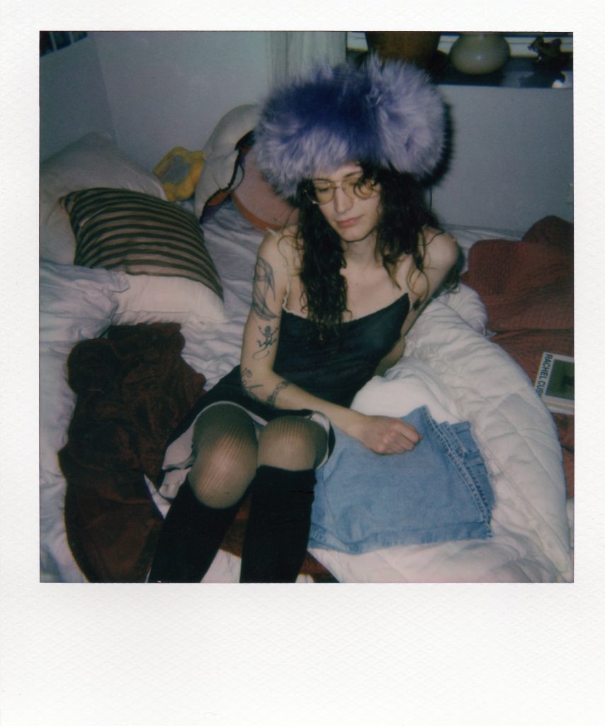 Image: Riley Yaxley, Avery on the Bed, 2025. A woman sits on a bed amid a tangle of blankets and pillows. She is wearing a black slip dress and a lavender fur hat. 