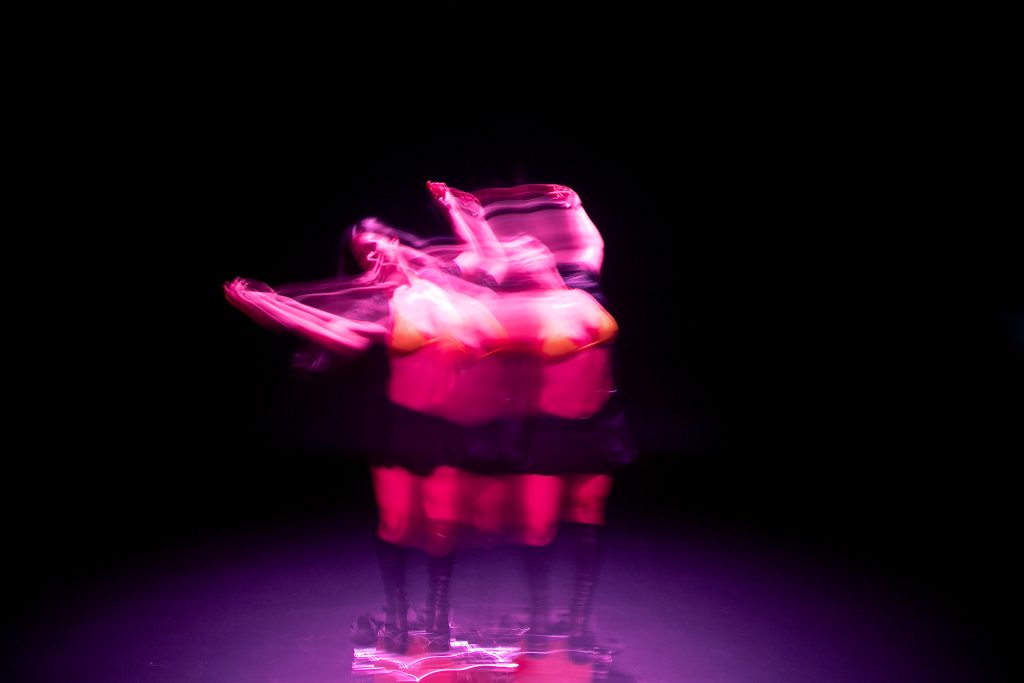 Image: Blurred double-image of choreographer D Hunter dancing underneath a pink spotlight during rehearsal at the Red Eye theater. Photo by Wordsworth Musinguzi.