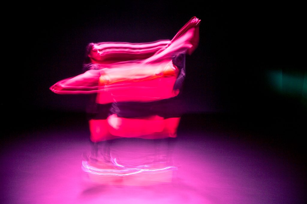 Image: Blurred double-image of choreographer D Hunter dancing underneath a pink spotlight during rehearsal at the Red Eye theater. Photo by Wordsworth Musinguzi.