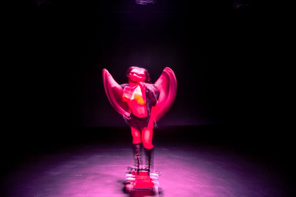 Image: Blurred image of D Hunter in-motion under pink spotlight light while standing on a step stool during rehearsal at the Red Eye. Photo by Wordsworth Musinguzi.