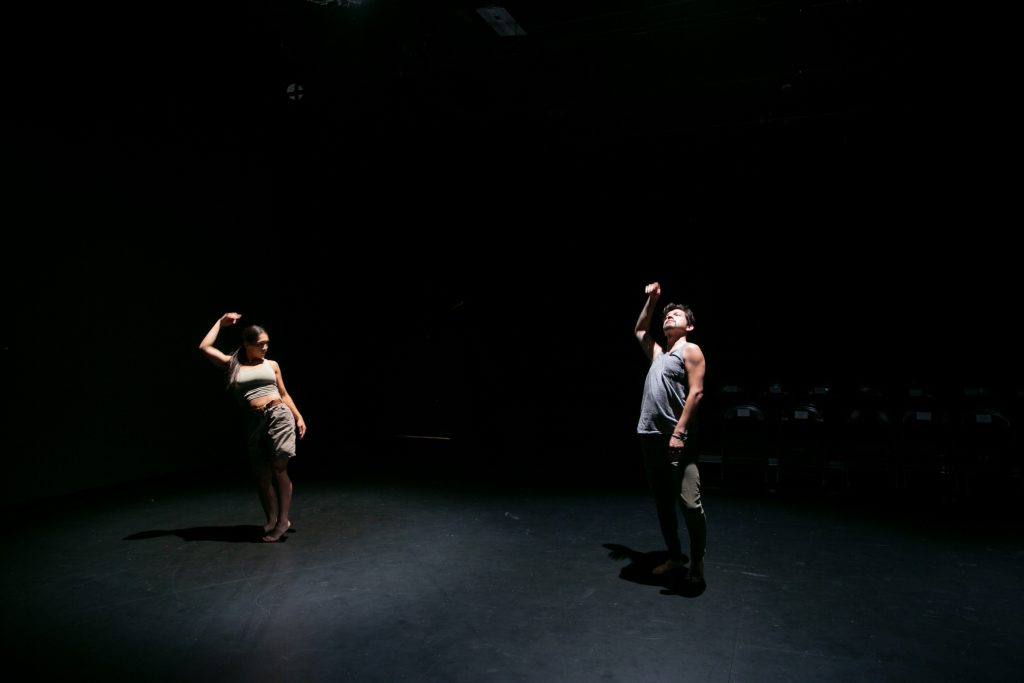 Image: D Hunter (left) and José A. Luis pose underneath spotlights in a darkened rehearsal space at the Red Eye in Minneapolis, MN. Photo by Wordsworth Musinguzi.
