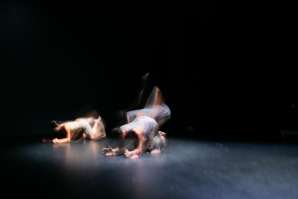 Image: Underneath a spotlight, D Hunter (left) and José A. Luis dancing with their elbows and knees on a black floor during rehearsal at the Red Eye in Minneapolis, MN. Photo by Wordsworth Musinguzi.