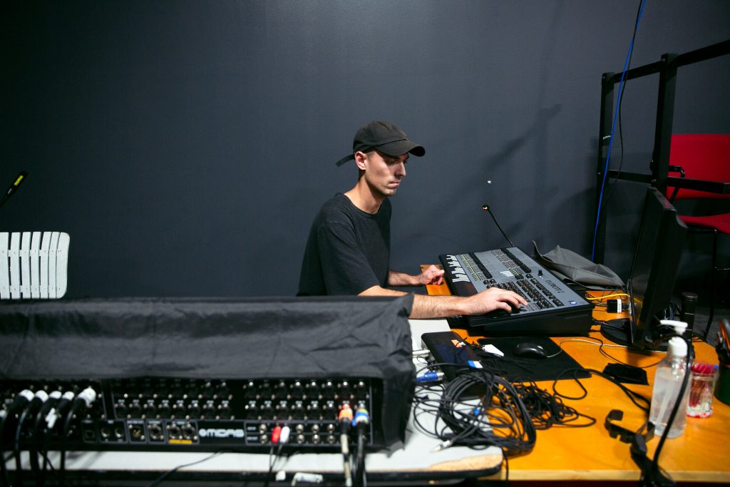 Image: más y more Stage Hand/Production Assistant, Connor Berkompas, sits behind the soundboard and adjusts the audio levels in preparation for rehearsal. Photo by Wordsworth Musinguzi.