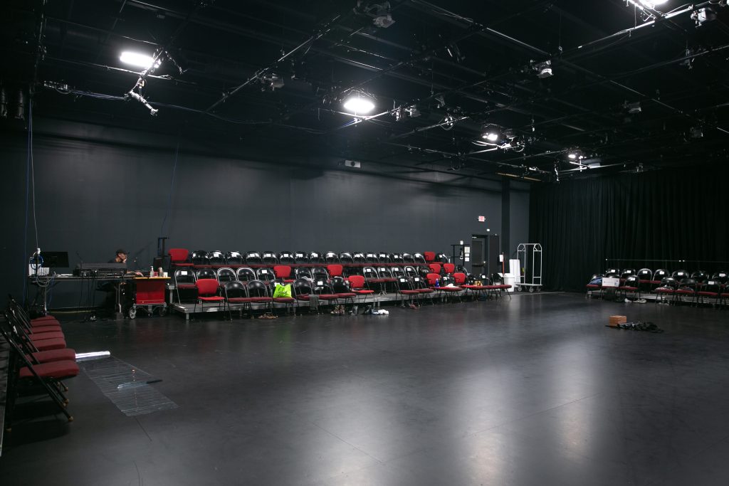 Image: A large black box theater rehearsal space is lined with rows of foldable chairs. A sound technician works behind a soundboard system near two rows of chairs at the far-left side of the room. Photo by Wordsworth Musinguzi.