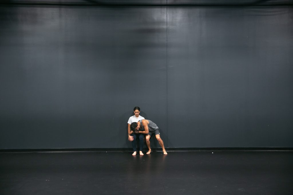 Image: D Hunter and José A. Luis embrace while balancing their bodies against a gray wall during rehearsal at the Red Eye theater. Photo by Wordsworth Musinguzi.