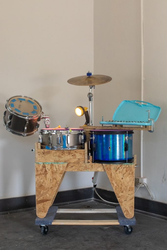 Image: A homemade drum set. Drums are inlaid on wood and set upon a furniture dolly. In the center of the drum set is a flashlight. On the right side is a blue xylophone. Image by Ruby Que.