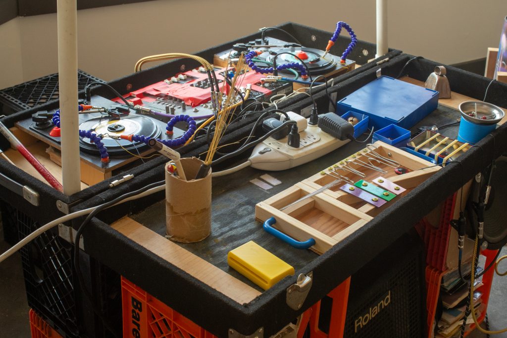 Image: Homemade DJ station which includes a crate seat and other homemade instruments for plucking, strumming, and shaking. Rows of 45s are stacked in crates under the DJ table. Image by Ruby Que.
