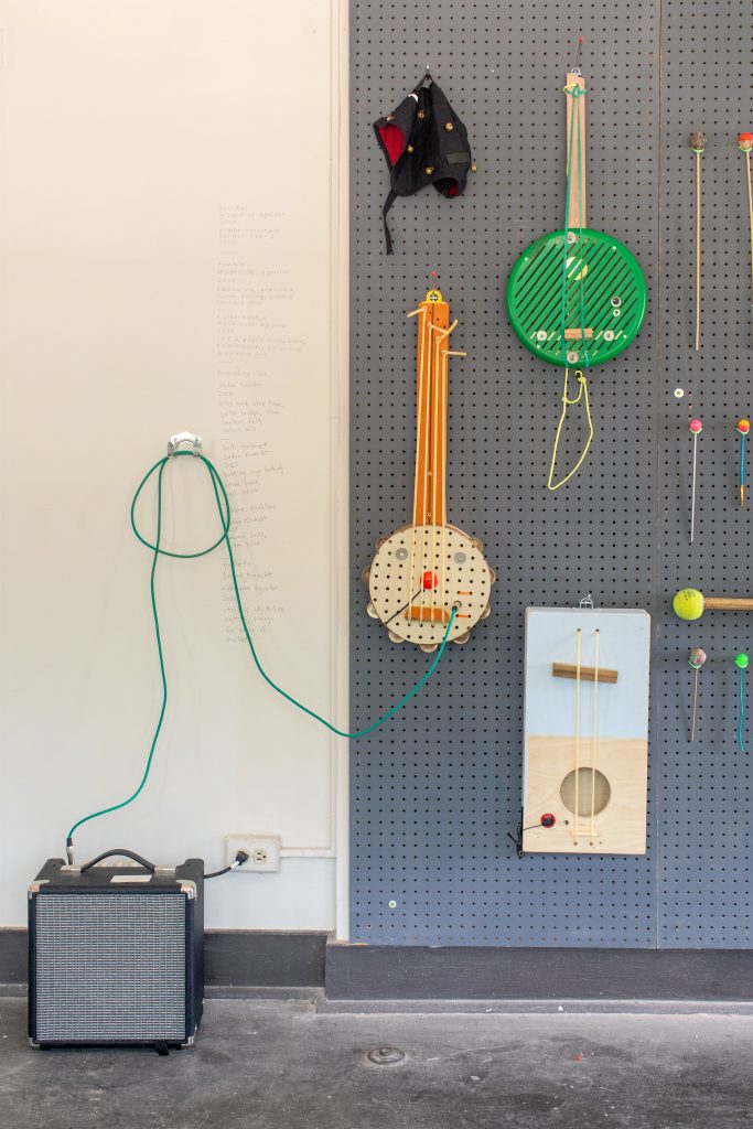 Image: Colorful homemade instruments hang on a grey pegboard wall. On the floor next sits an amp which is connected to a rubber banjo. Image by Ruby Que.