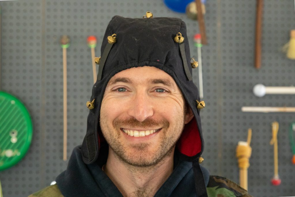 Image: Knecht smiles, front and center, wearing a homemade black cap with bells. Behind him is the pegboard wall of instruments he crafted with Aguilar. Image by Ruby Que.