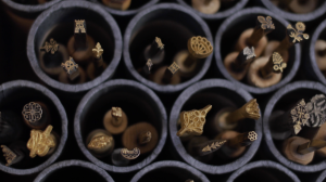 Image: a close up on the openings of black tubes stacked in a hive-like rows. Inside each tube are small golden, bronzed, and silver stamps of various shapes. Some appear to be flowers, one is a castle tower, another is a skull and crossbones, others are abstract shapes. Image by Ian Carstens.