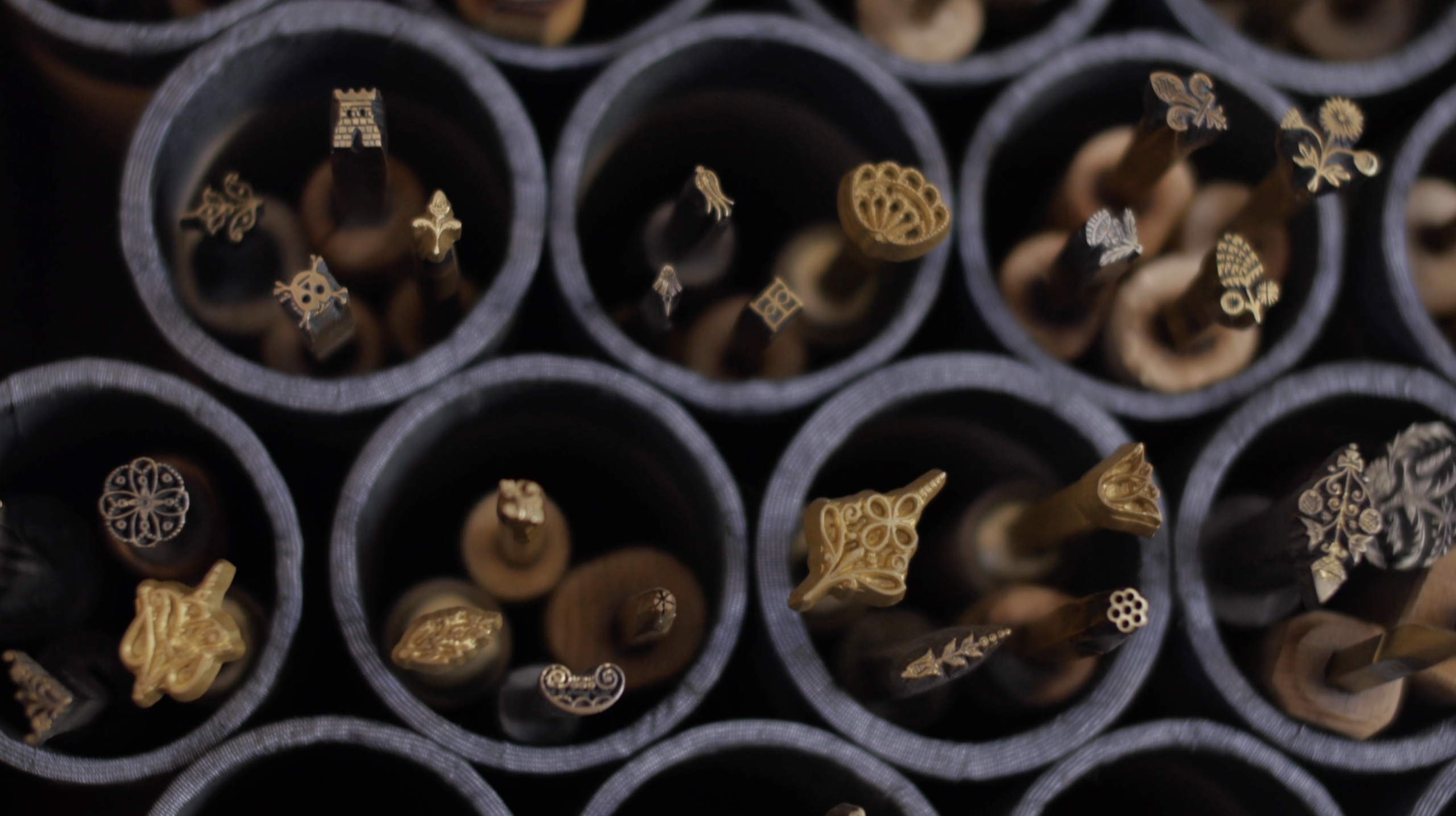Image: a close up on the openings of black tubes stacked in a hive-like rows. Inside each tube are small golden, bronzed, and silver stamps of various shapes. Some appear to be flowers, one is a castle tower, another is a skull and crossbones, others are abstract shapes. Image by Ian Carstens.