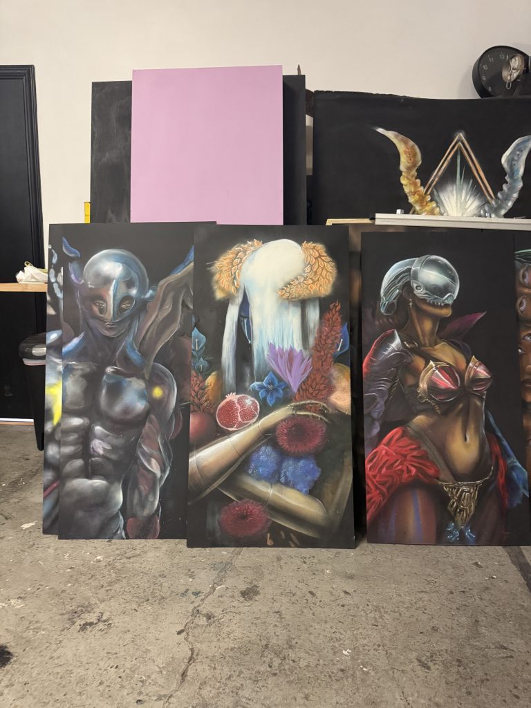 Image: a trio of canvas paintings of various mystical characters. The far left is an angelic man in armor and the far right is an armored woman. In the middle is Emma Stone's original character larger and cropped to their bust up. Image by Claire Stemen.