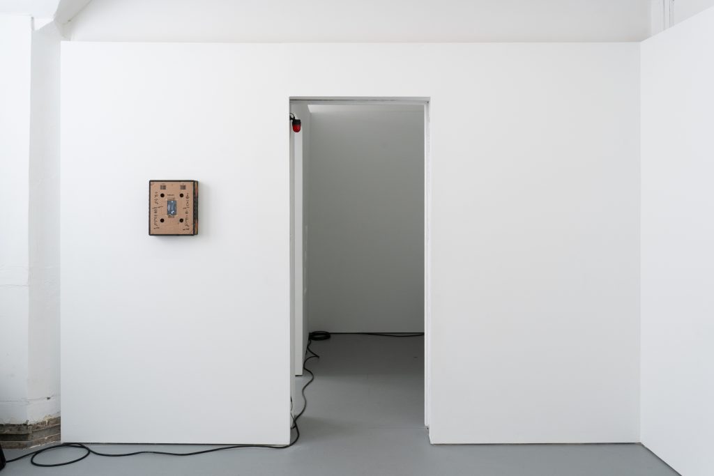 Image: Installation view of the exhibition with a doorway in the center. Left of the door, Speech Assistance Machine (Leonard Cohen’s Democracy, 1992) hangs on the wall. A wire runs on the ground between the rooms. Image courtesy of Letters to Nora.