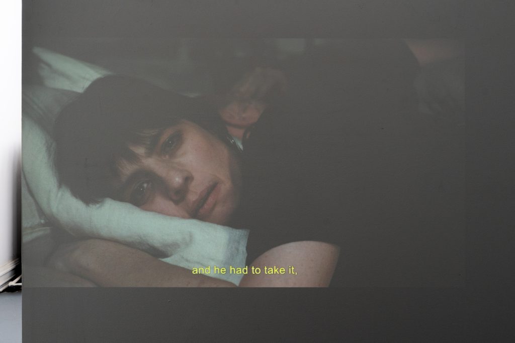Image: A projection on the wall from the video A Thousand Telephones (scenes 1-5) with musical arrangement by Anna Arrobas and Shane Hoy. A projection is a close-up of a person lying in a bed with their head resting on a pillow. A caption at the bottom of the projection reads "and he had to take it." Image courtesy of Letters to Nora.