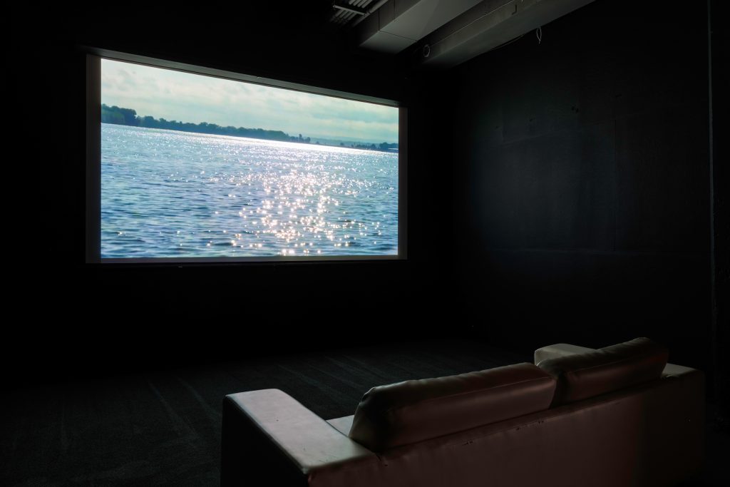 Image: A still shot of the surface of a lake on a partly cloudy day in Asad Raza's Delegation, 2022, video playing in the theater room in front of a brown couch at Bemis Center. Courtesy of the artist  Image courtesy Bemis Center for Contemporary Arts. Photo by Colin Conces. 