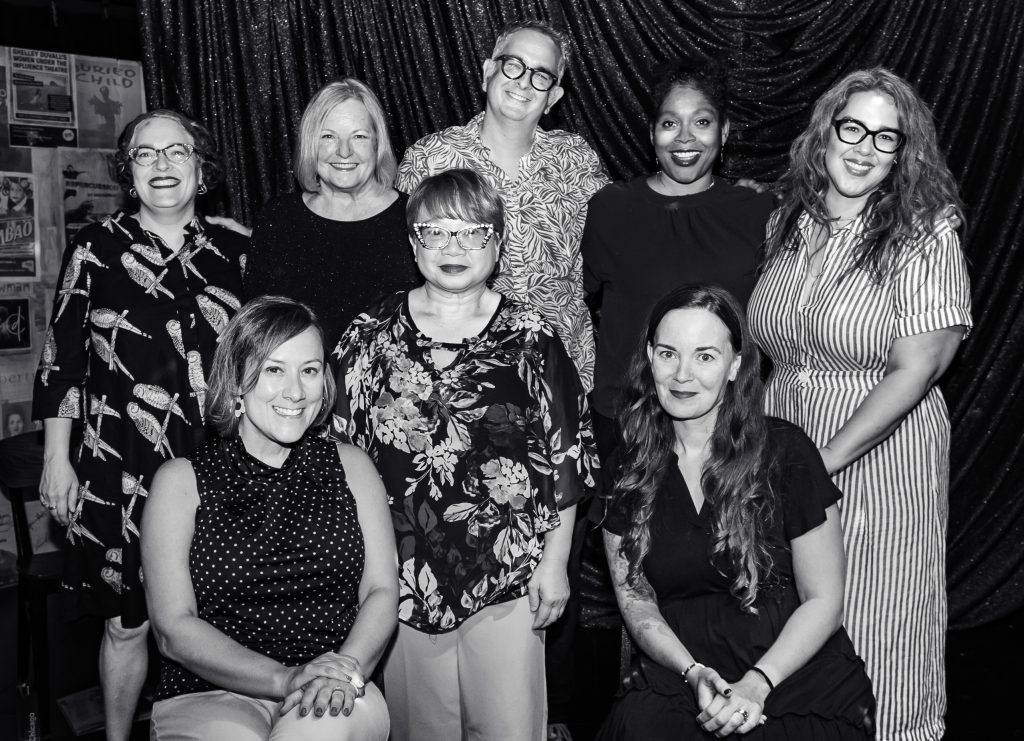 A posed black and white photo of a group of 8 smiling people. Maggie in a dark polka dot top is seated at the lower left.