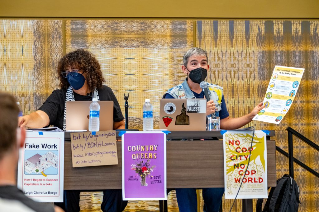 Two panelists are seated at a table with laptops before them. There are various posters with political slogans affixed to the table. The panelist on the right is speaking into the microphone while holding a flyer titled "How to Clean Up Copaganda in Ten Simple Steps."