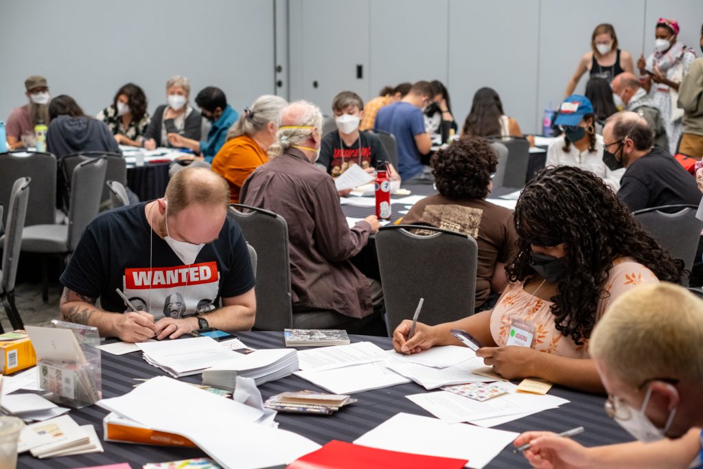 A large group of conference attendees are seated around round tables while writing. Various utensils cover the surface of the tables.