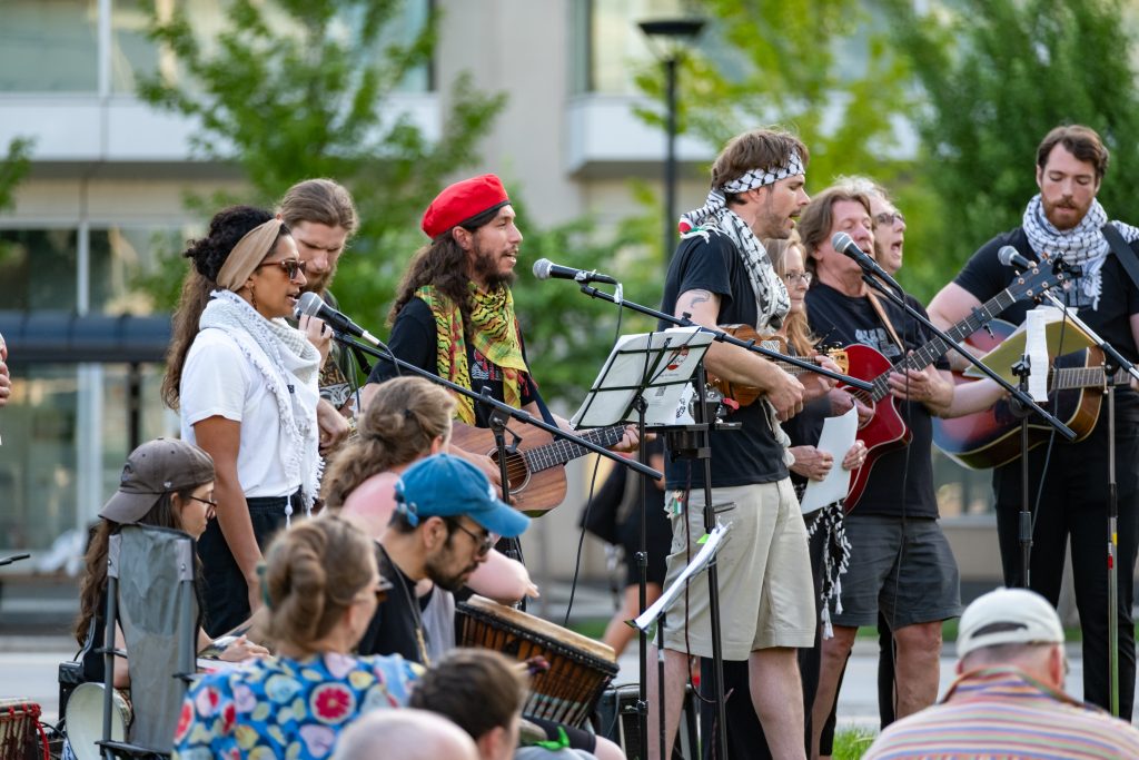 A group of musicians perform outside for an audience. Several are singing into microphones and playing acoustic guitars. A few of the performers are wearing keffiyehs.