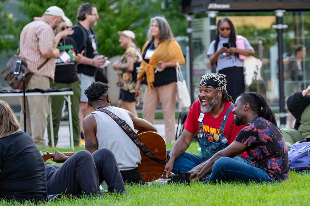 A small group of conference attendees hang out on the lawn. One of the attendees is playing a guitar while the other two laugh with glee. Other attendees are standing in the background.