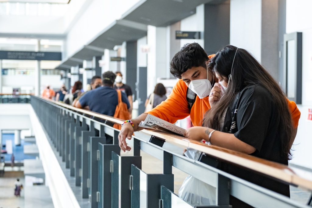 Two attendees wearing masks consult the conference program while leaning against a bannister. Other attendees are visible in the background, heading to and from conference sessions.