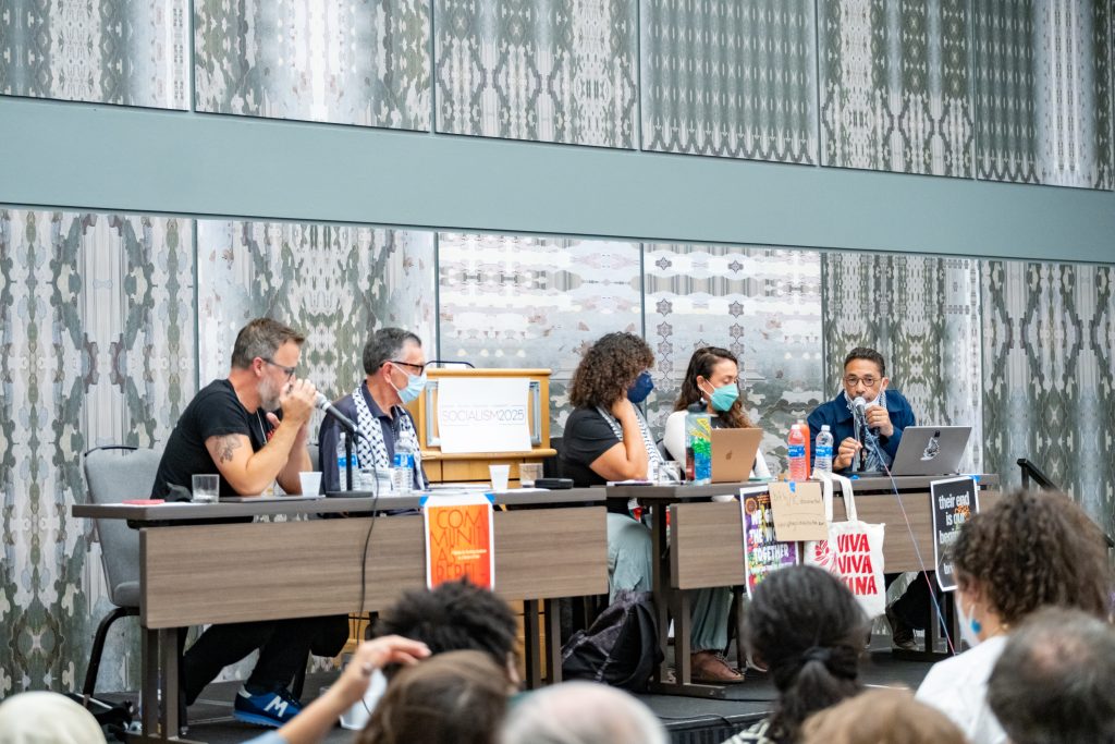 Five panelists are seated behind a table before a crowd. The panelist at the far end of the table is speaking into a microphone. There are various posters with political slogans affixed to the table.
