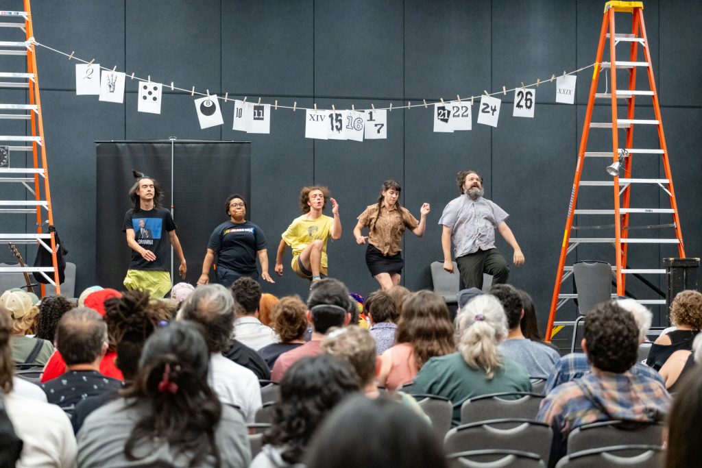 A crowd watches five performers onstage. The performers appear to be running. There are two orange ladders on either side of the performers with a series of numbers strung between them.
