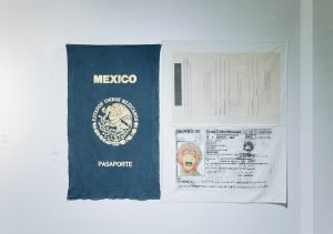 Image: Installation view from We Built This: Making Through Migration. Pasaporte de Abelardo Montoya by Keny De La Peña is a large textile artwork resembling a Mexican passport. The left panel shows the gold emblem and text “MEXICO PASAPORTE,” while the right panels recreate passport pages one blank, the other featuring a Muppet-like character as the ID photo. Image courtesy of Zhou B Art Center.