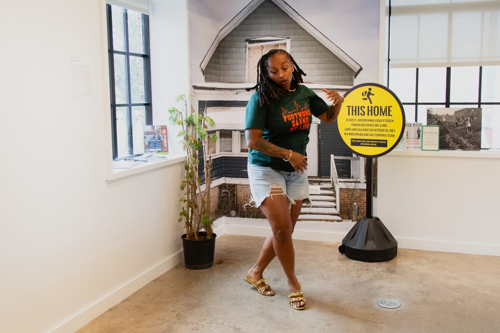 Dr. ShaDawn Battle breaks out into dance inside of the National Public Housing Museum. Behind her is an image of a small house that is wrapped around the corner of a wall. A tall plant in a black pot is on the left side of the image, a large yellow sign with small text is on the right side of the image, while various flyers, books, and paper materials rest on the window sills of the room. Image by Joshua Clay Johnson.