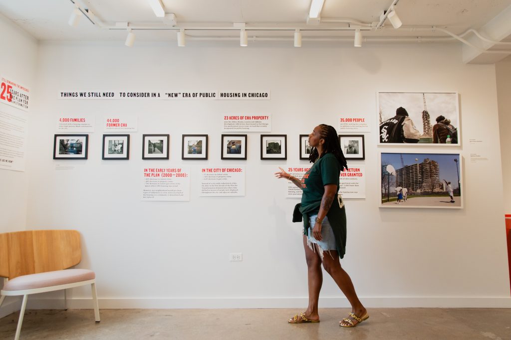 Dr. ShaDawn Battle looks at the exhibit "Things We Still Need to Consider in a "New"Era of Public Housing in Chicago". On a white gallery wall hang eight images. They are displayed in black frames and hung horizontally as if on a timeline. The images are too far away to see what they are of. At the far right are two much larger images in white frames. The top one features two people looking away from camera and the bottom features people playing on an outdoor basketball court. Amongst the images are boxes of black and red text. Image by Joshua Clay Johnson.